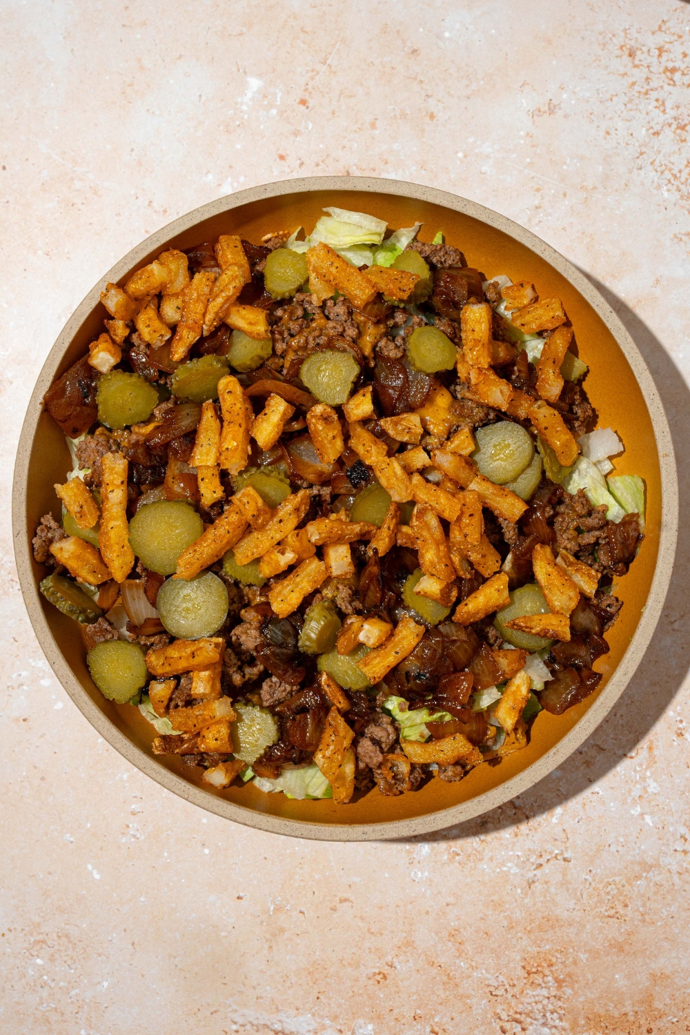 An orange plate with shredded lettuce and ground burger meat with melted cheese, french fries, pickles, and caramelized onions. The plate is on a tan counter.