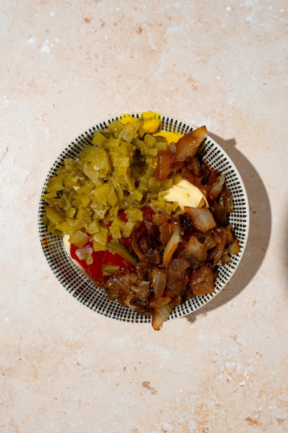 A plate with caramelized onions, mayo, ketchup, and relish on a tan counter.