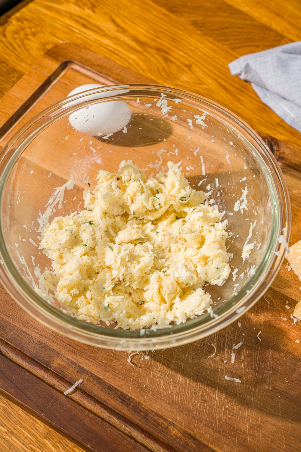 A glass bowl with a parmesan cheese ball mixture including parmesan cheese, egg, and seasonings. The bowl is on a wooden cutting board.