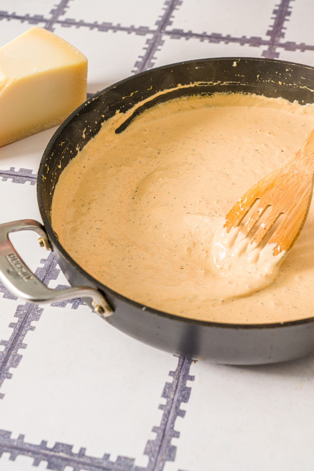 A skillet with a wooden spoon stirring a cajun pasta sauce. The skillet is on a tiled counter.