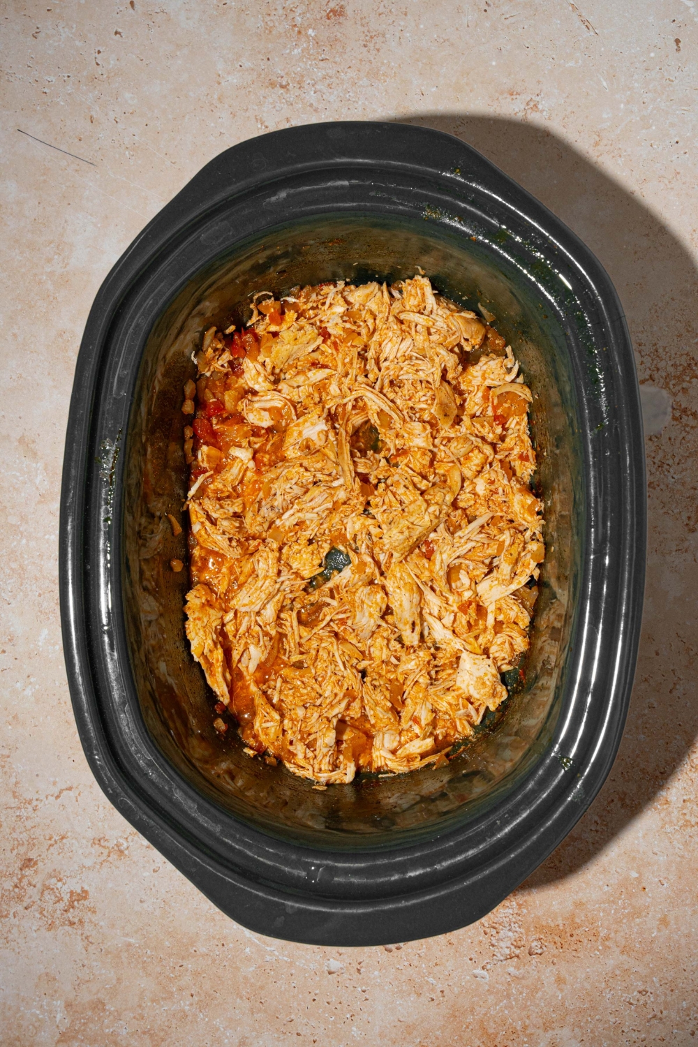 A slow cooker with cooked shredded chicken tossed in Buffalo sauce and seasonings. The slow cooker is on a tan counter.