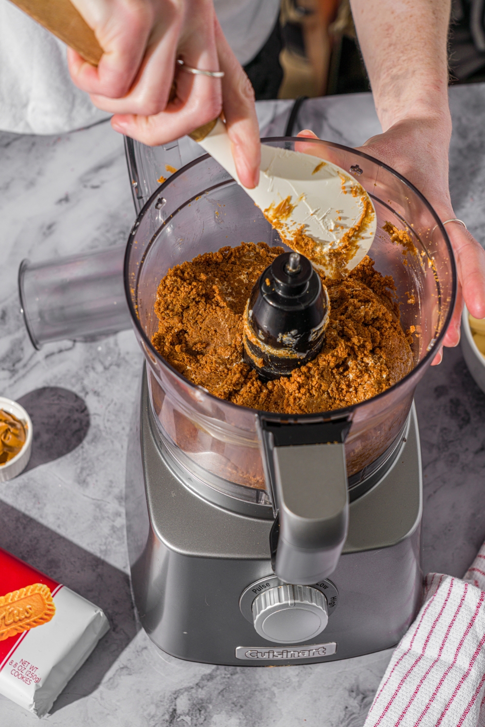 A food processor with crumbled Biscoff cookies. A spatula is pushing the crumbs down. The food processor is on a marble counter.