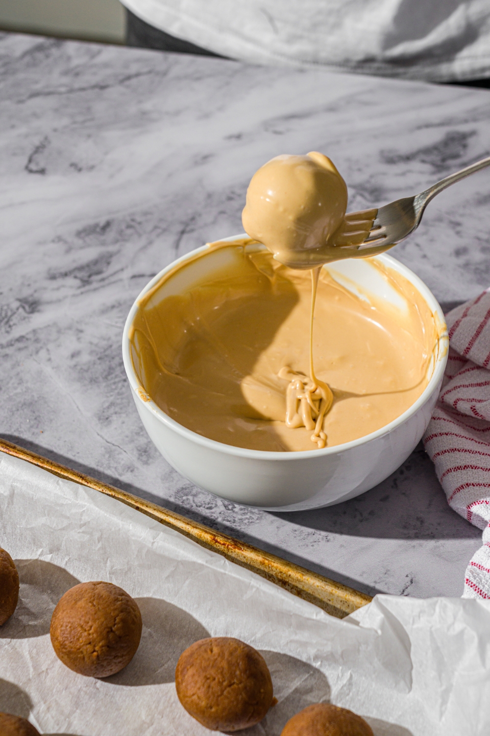 A white bowl of melted white chocolate with a fork dipping a Biscoff truffle into the chocolate. The bowl is on a marble counter with a baking sheet of truffles.