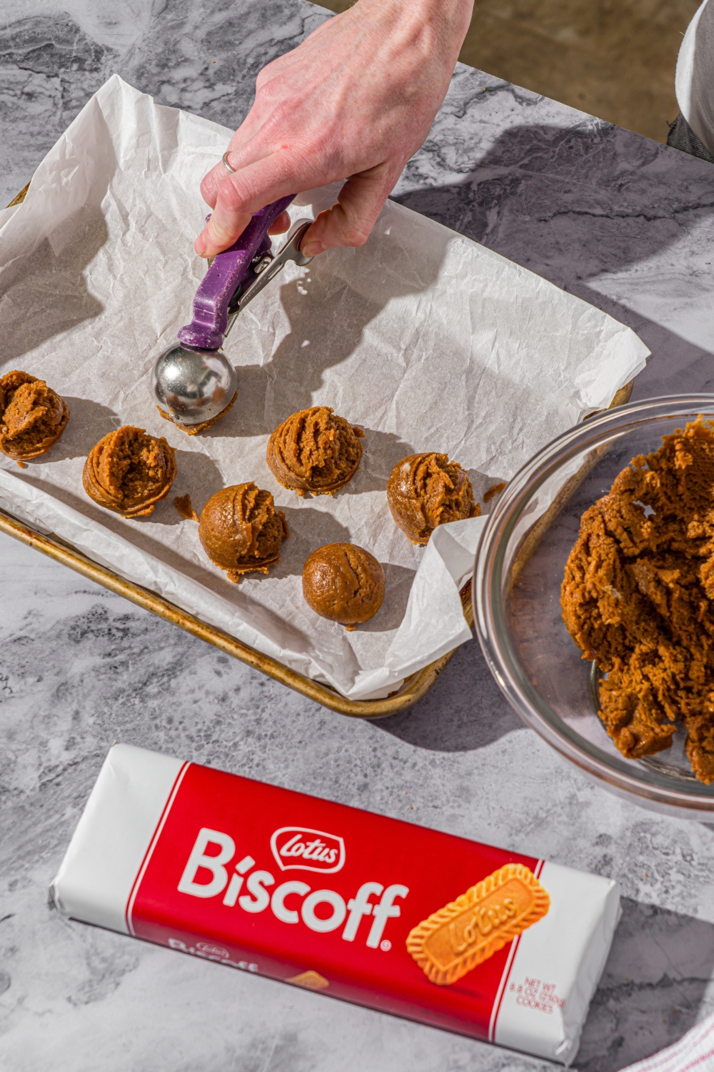 A Biscoff truffle mixture being scooped from a glass bowl onto a baking sheet lined with parchment paper. The sheet is on a marble counter.