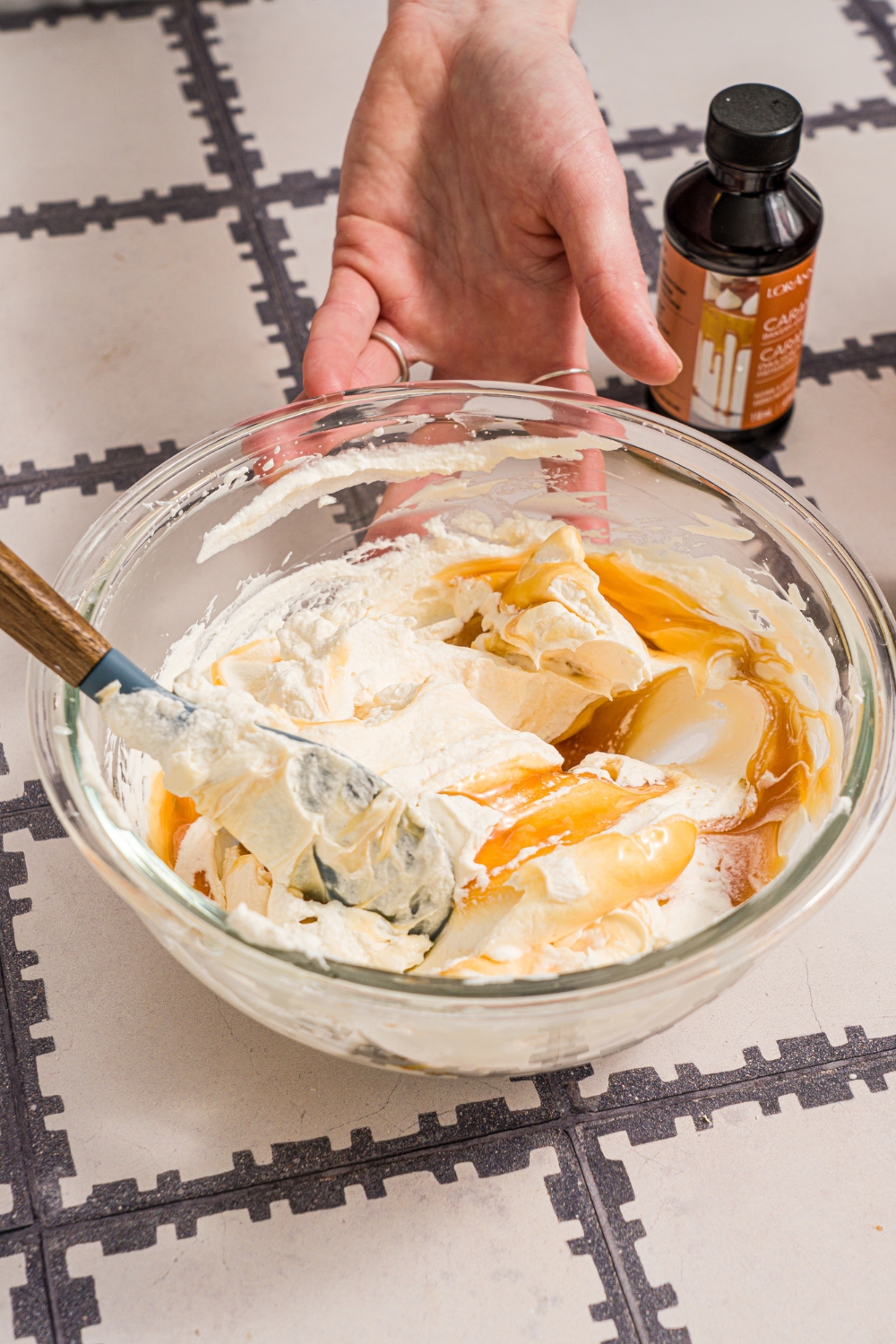 A large glass bowl with a spatula mixing butterscotch syrup with whipped cream. The bowl is on a tiled counter.