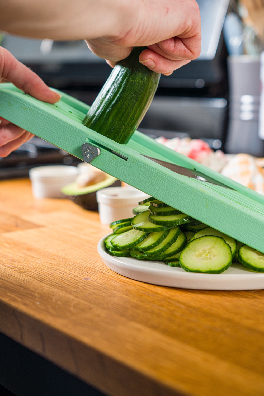A mandolin slicing a whole cucumber over a white plate on a wooden counter.