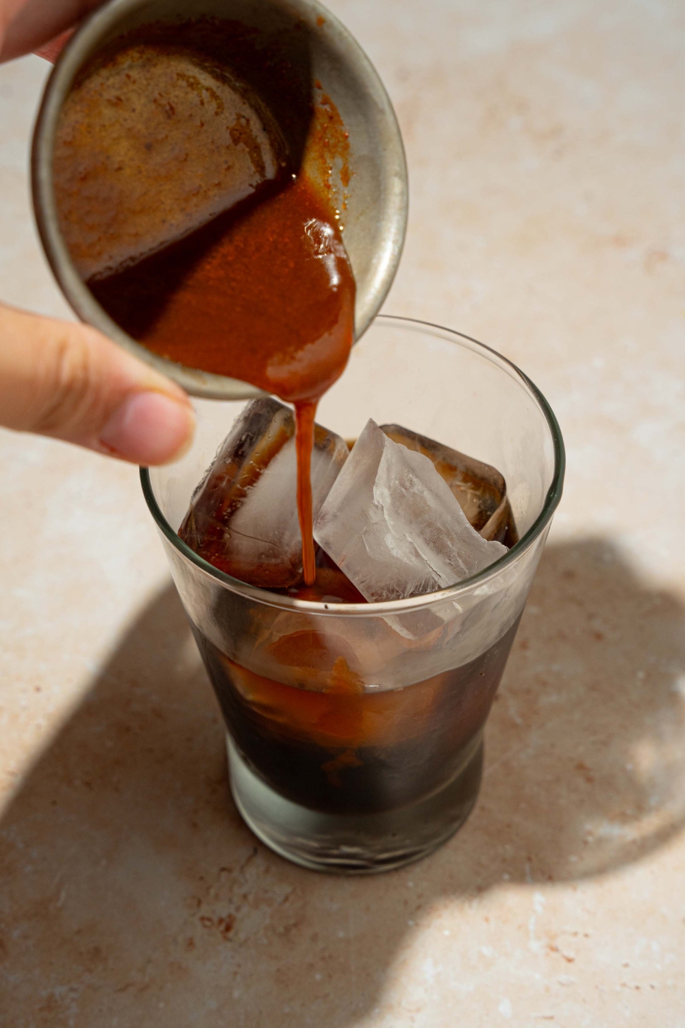 A glass tumbler with carrot cake syrup being added to an iced espresso. The glass is on a tan counter.