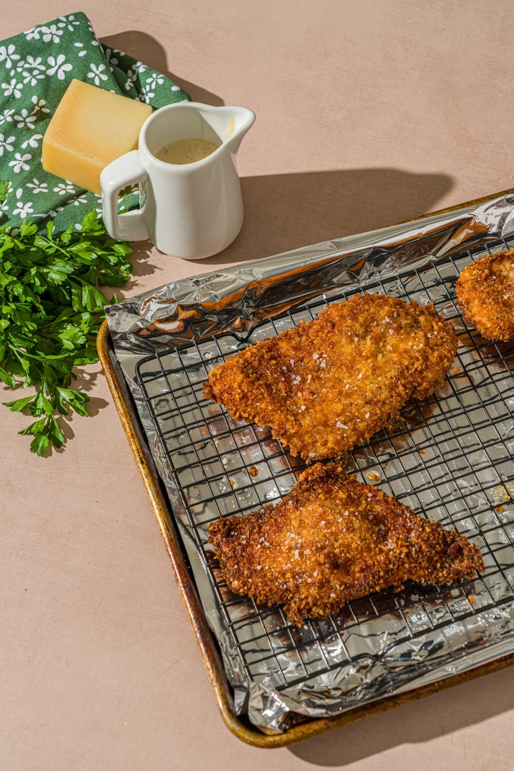 A baking sheet lined with foil with a rack on top. There are pieces of fried breaded cutlets on the rack. The sheet is on a tan counter with a creamer of dressing.