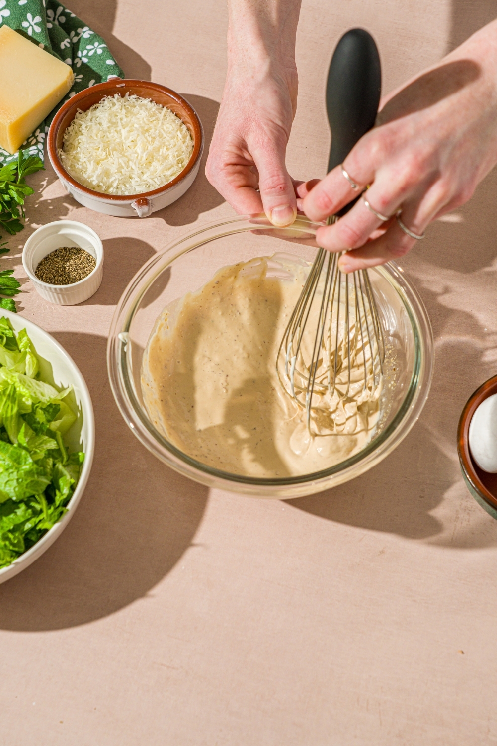 A glass bowl with a whisk mixing ingredients to make caesar dressing. The bowl is on a tan counter with bowls of ingredients.