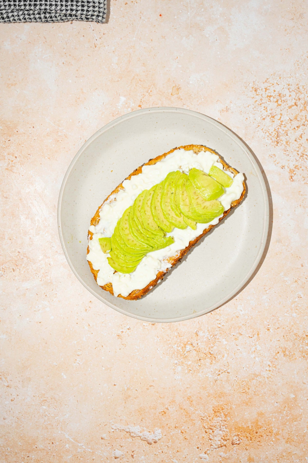 A white plate with a slice of toast layered with cottage cheese and avocado. The plate is on a tan counter.