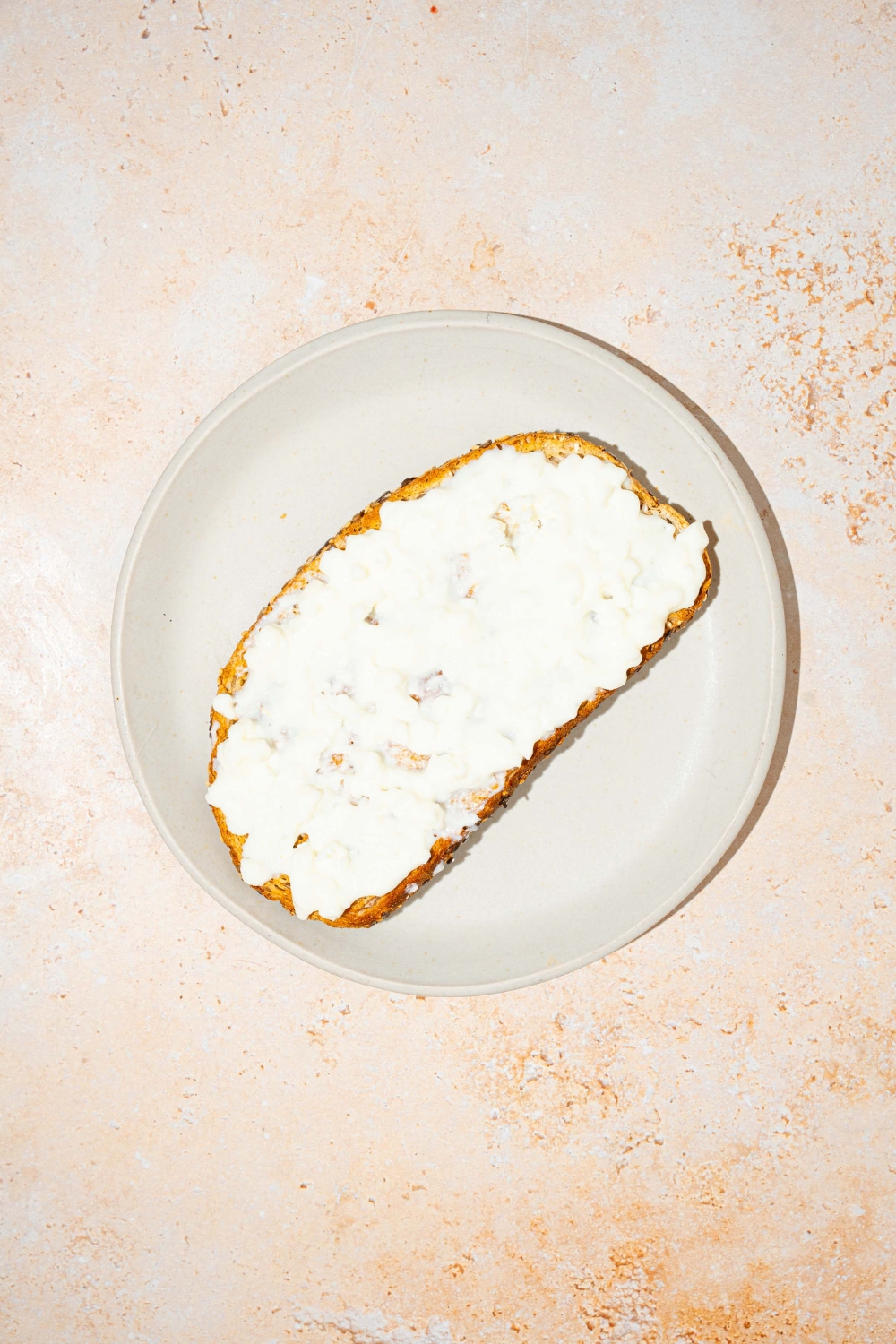 A white plate with a slice of toast with cottage cheese. The plate is on a tan counter.