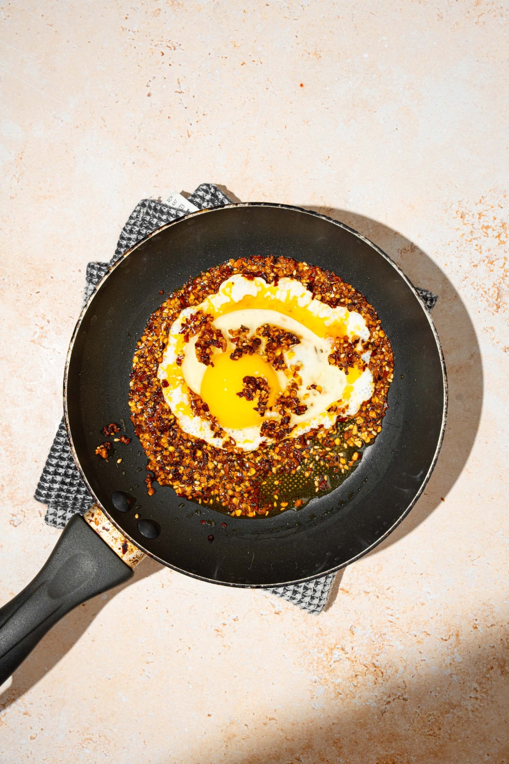 A skillet with an egg frying in chili crisp oil. The skillet is on a gray cloth napkin on a tan counter.