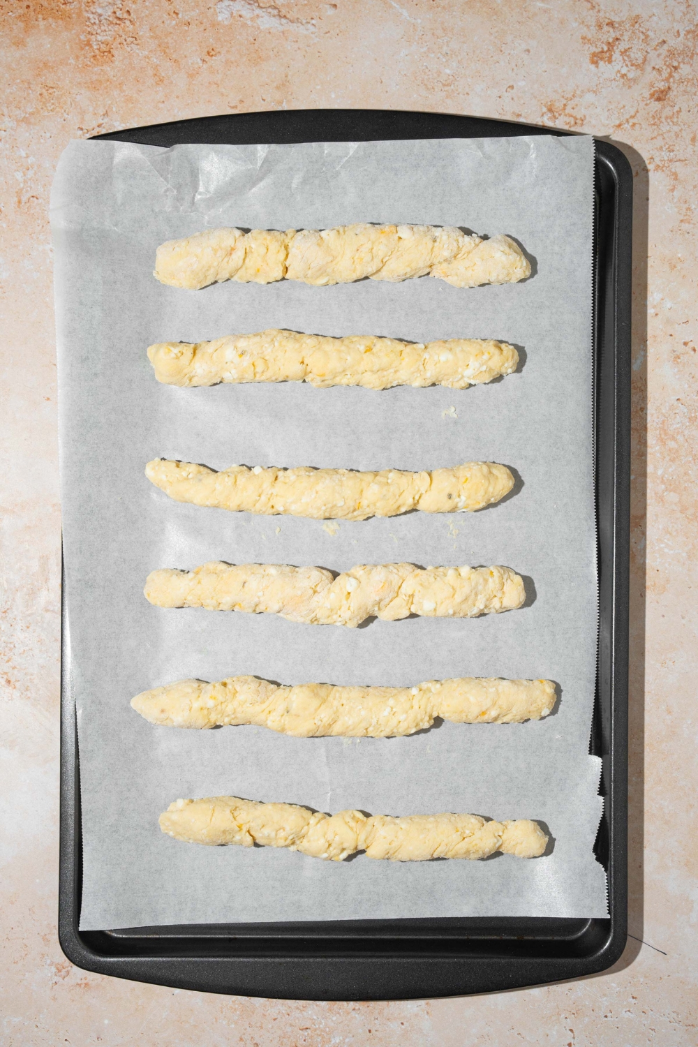 A baking sheet lined with parchment paper with several unbaked cottage cheese breadsticks. The sheet is on a tan counter.