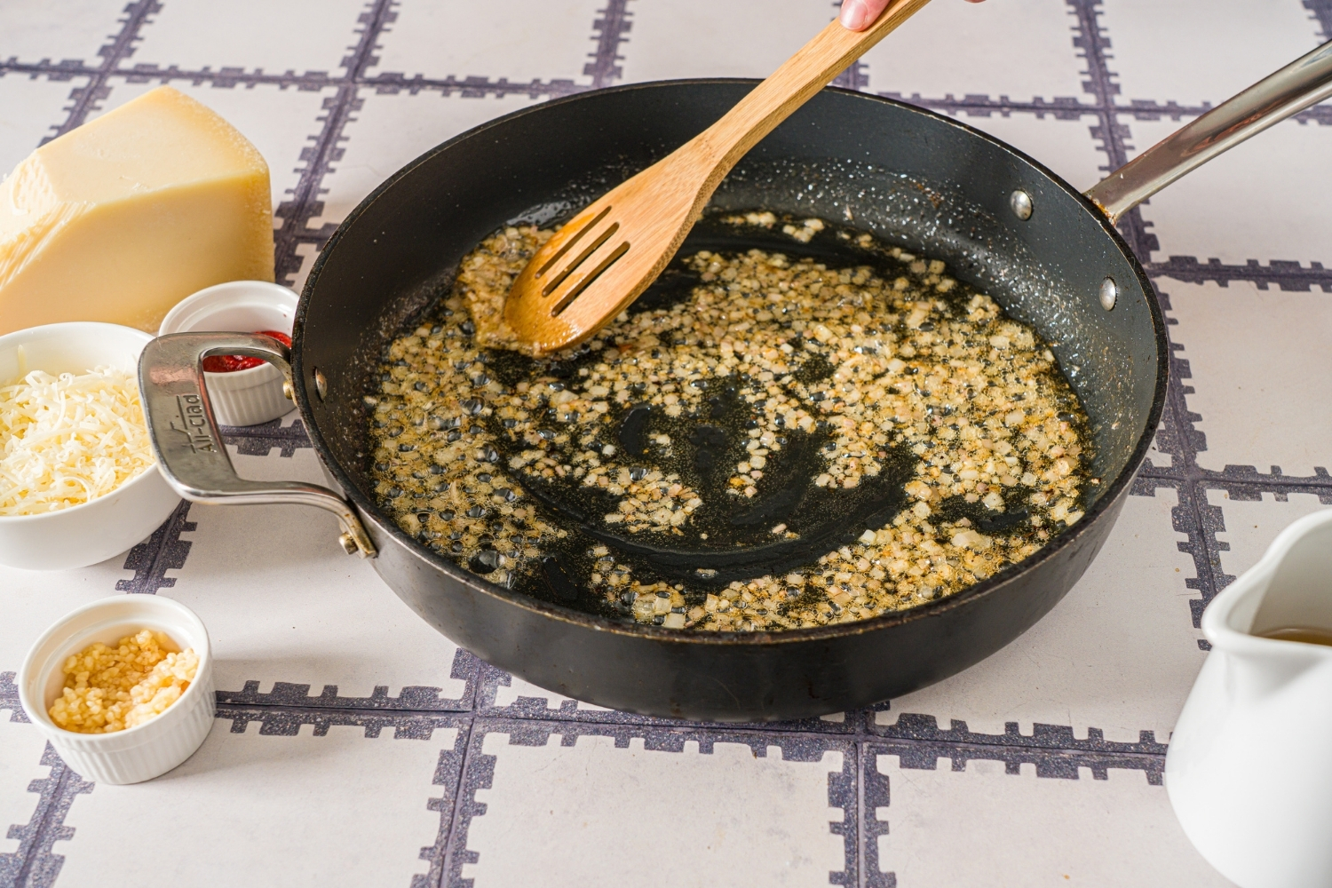 A skillet with a wooden spoon stirring garlic and shallots sautéing in oil. The skillet is on a tiled counter with small bowls of ingredients.