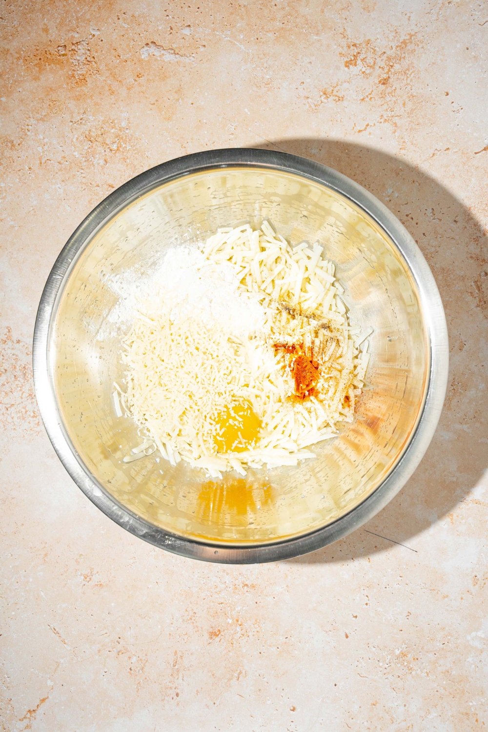 A large bowl with parmesan hashbrown ingredients including shredded potatoes, shredded parmesan cheese, egg, and seasonings. The bowl is on a tan counter.