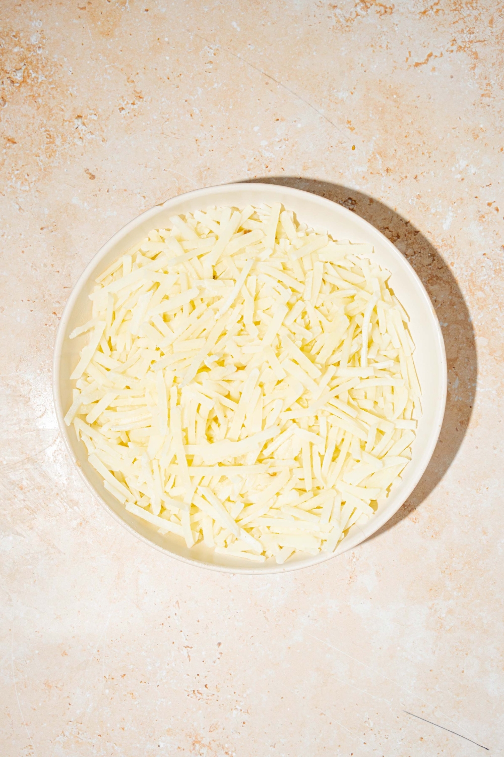 A white bowl with shredded potatoes. The bowl is on a tan counter.