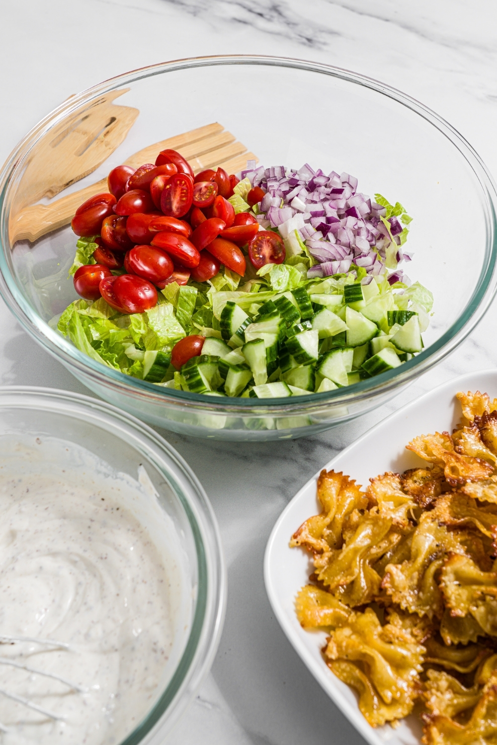 A glass bowl with ingredients to make salad including shredded lettuce, tomatoes, cucumbers, and red onion. The bowl is on a marble counter with a bowl of crispy pasta, a bowl of yogurt dressing, and two salad tongs.