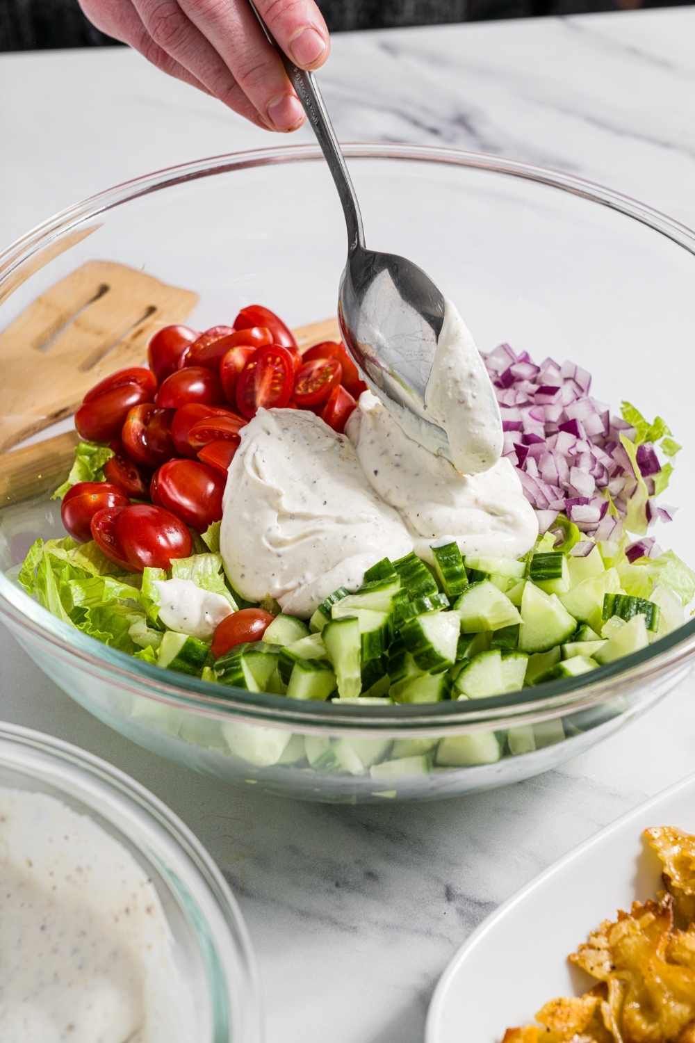A glass bowl with ingredients to make salad including shredded lettuce, tomatoes, cucumbers, and red onion. A spoon is pouring yogurt dressing over the vegetables. The bowl is on a marble counter.
