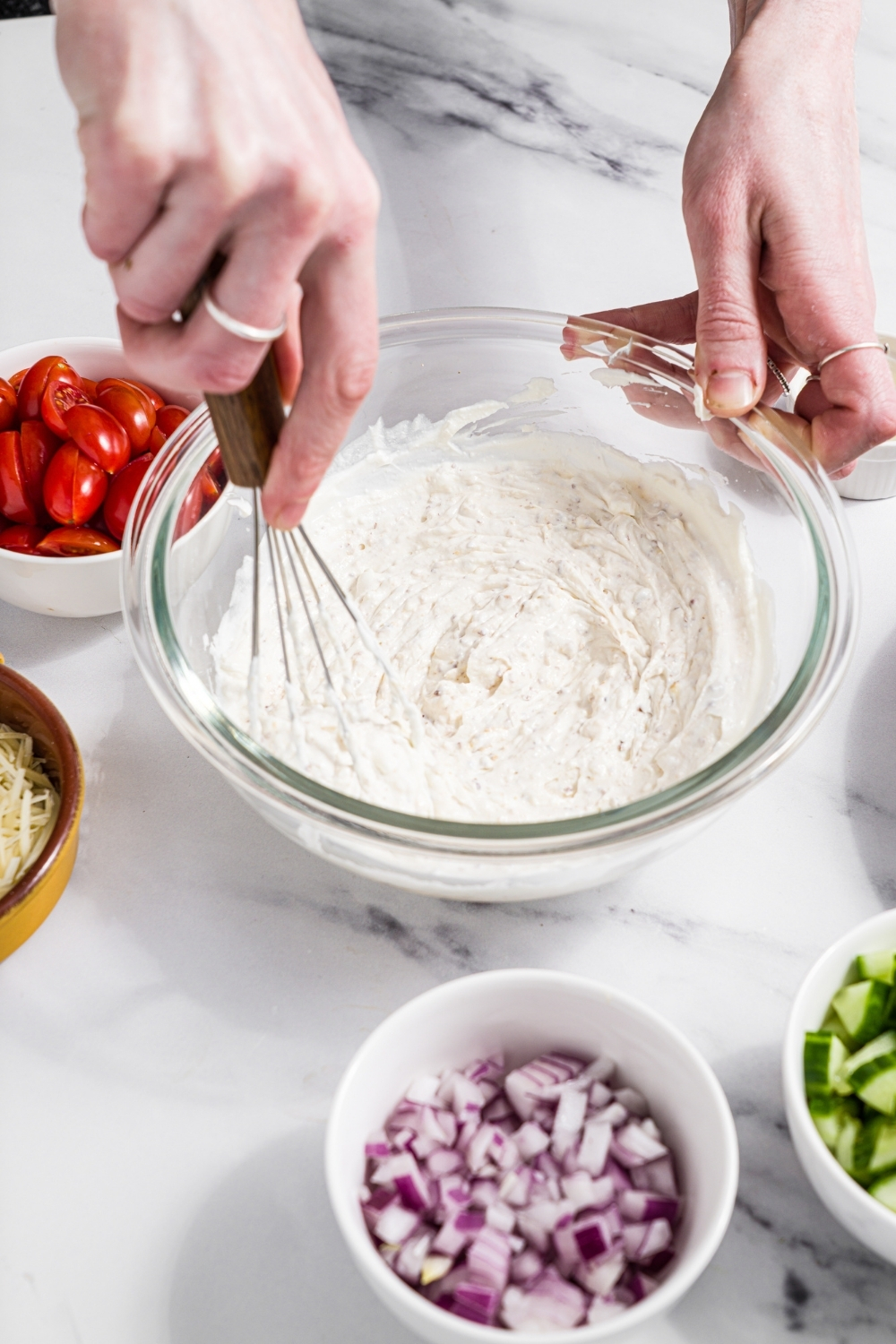 A glass bowl with a whisk mixing ingredients to make yogurt dressing including yogurt, sour cream, and seasonings. The bowl is on a marble counter with small bowls of salad ingredients.