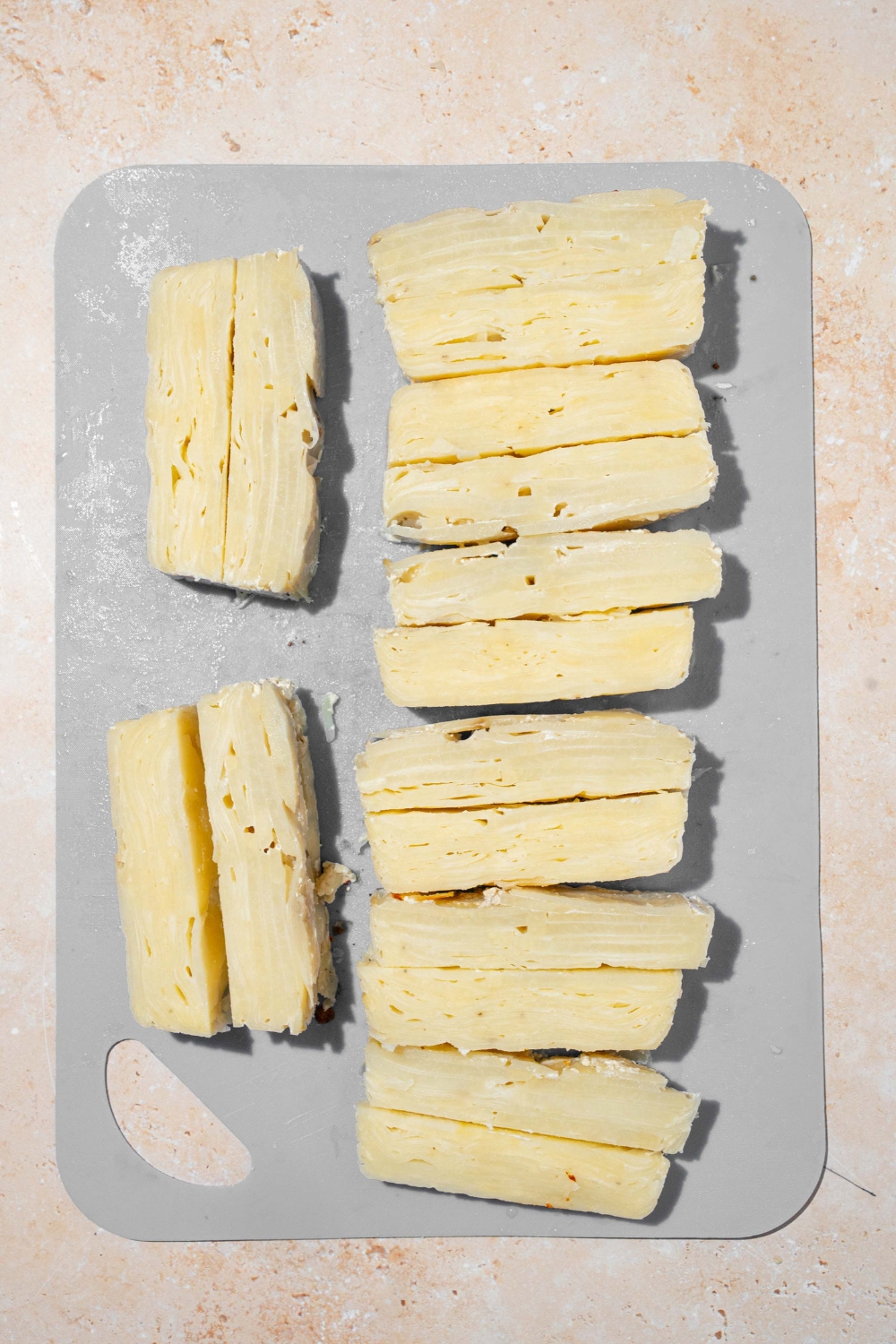 A cutting board with several pieces of sliced chilled potato pave. The cutting board is on tan counter.