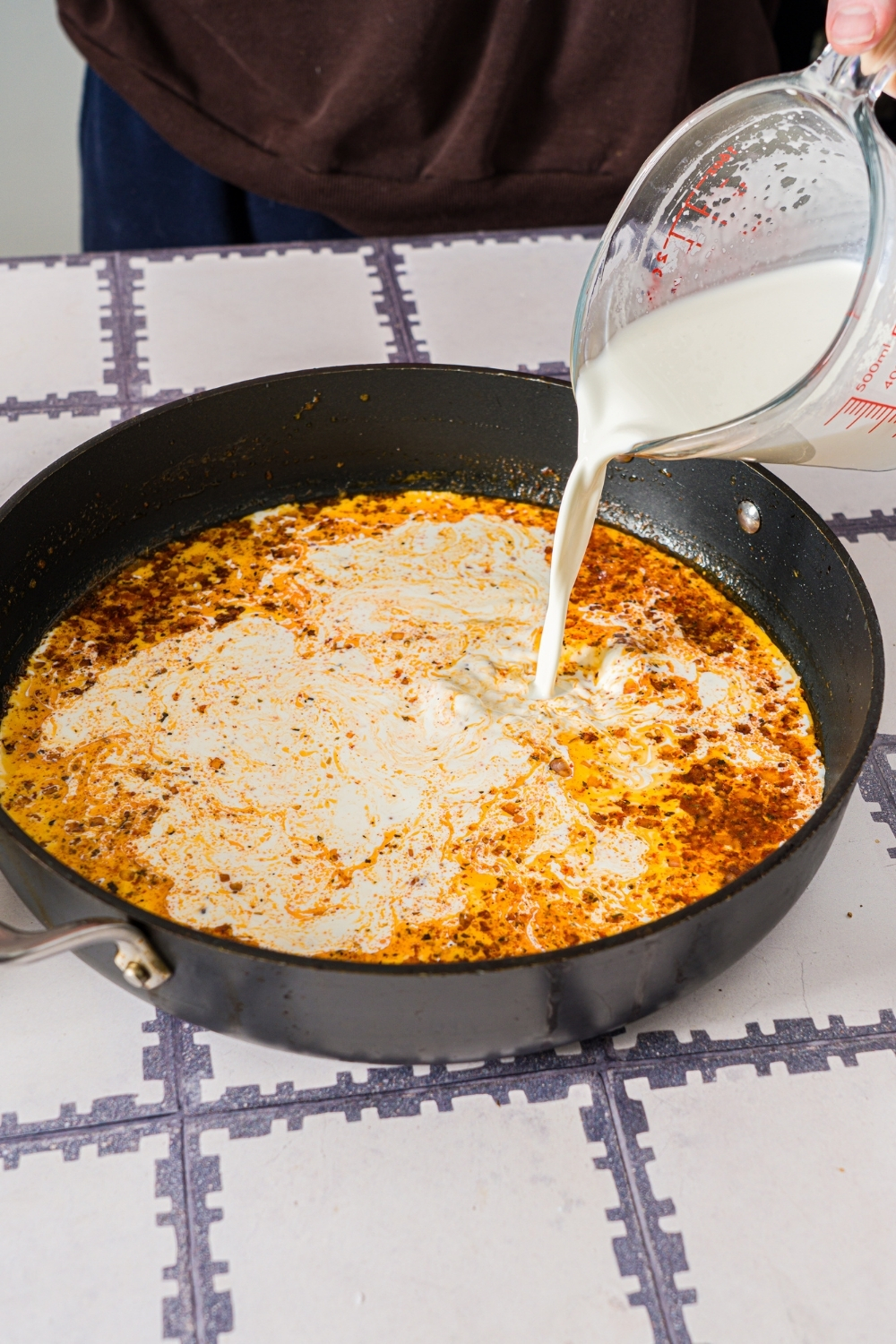 A skillet with heavy cream being added to a cajun sauce base. The skillet is on a tiled counter.