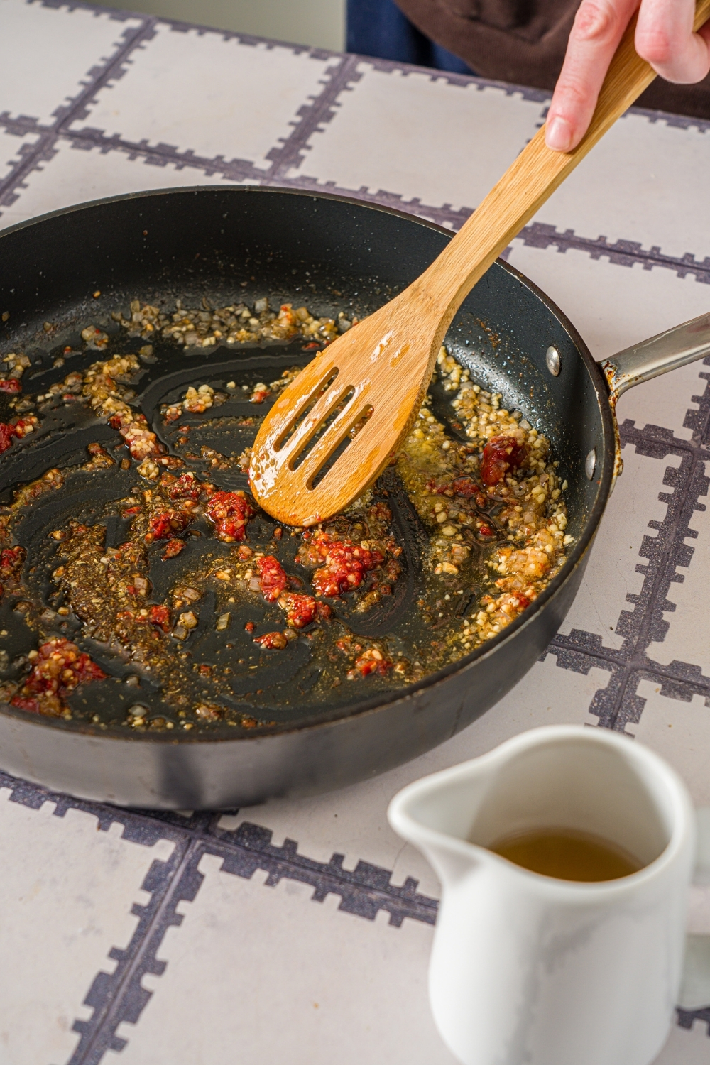 A skillet with a wooden spoon stirring garlic, tomato paste, and seasonings sautéing in oil. The skillet is on a tiled counter.