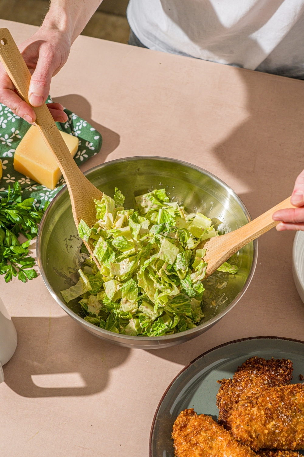 A large bowl with a pair of salad tongs tossing shredded romaine with caesar dressing. The bowl is on a tan counter.
