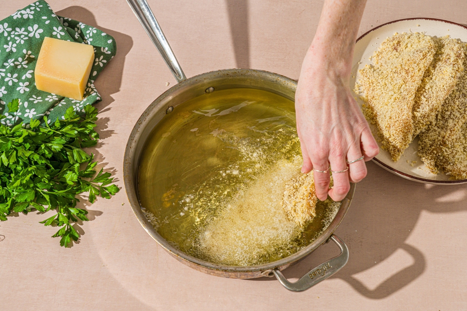 A breaded chicken cutlet being added to a skillet with hot oil. The skillet is on a tan counter with a plate of breaded cutlets.