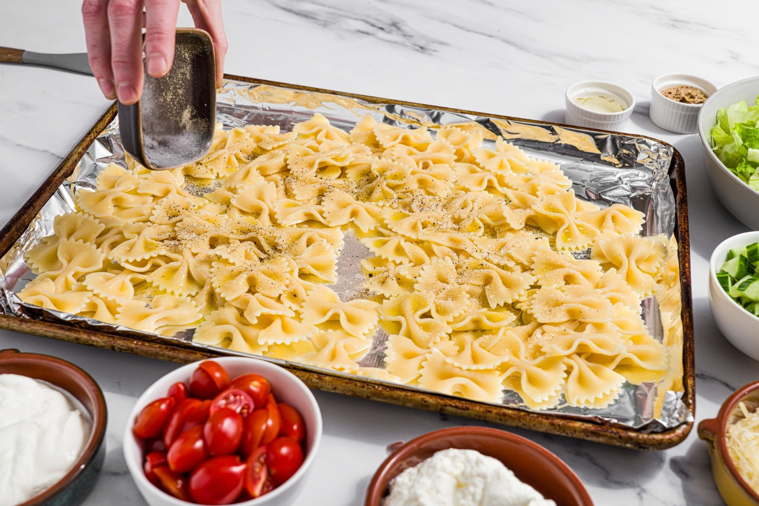 A baking sheet lined with foil with cooked bowtie pasta. There is a bowl of seasoning being poured over the pasta. The sheet is on a marble counter with small bowls of ingredients.