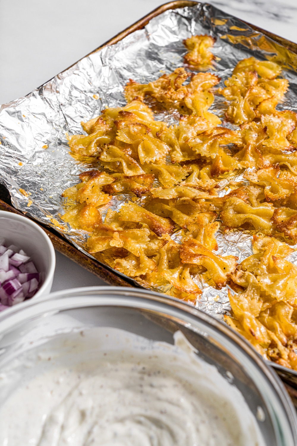 A baking sheet lined with foil with fried bowtie pasta. The sheet is on a marble counter with a bowl of yogurt dressing.