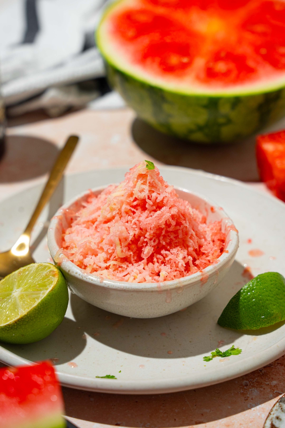 A small white bowl with watermelon shaved ice drizzled with honey. The bowl is on a white plate with a spoon and lime. The plate is on a tan counter with a watermelon.