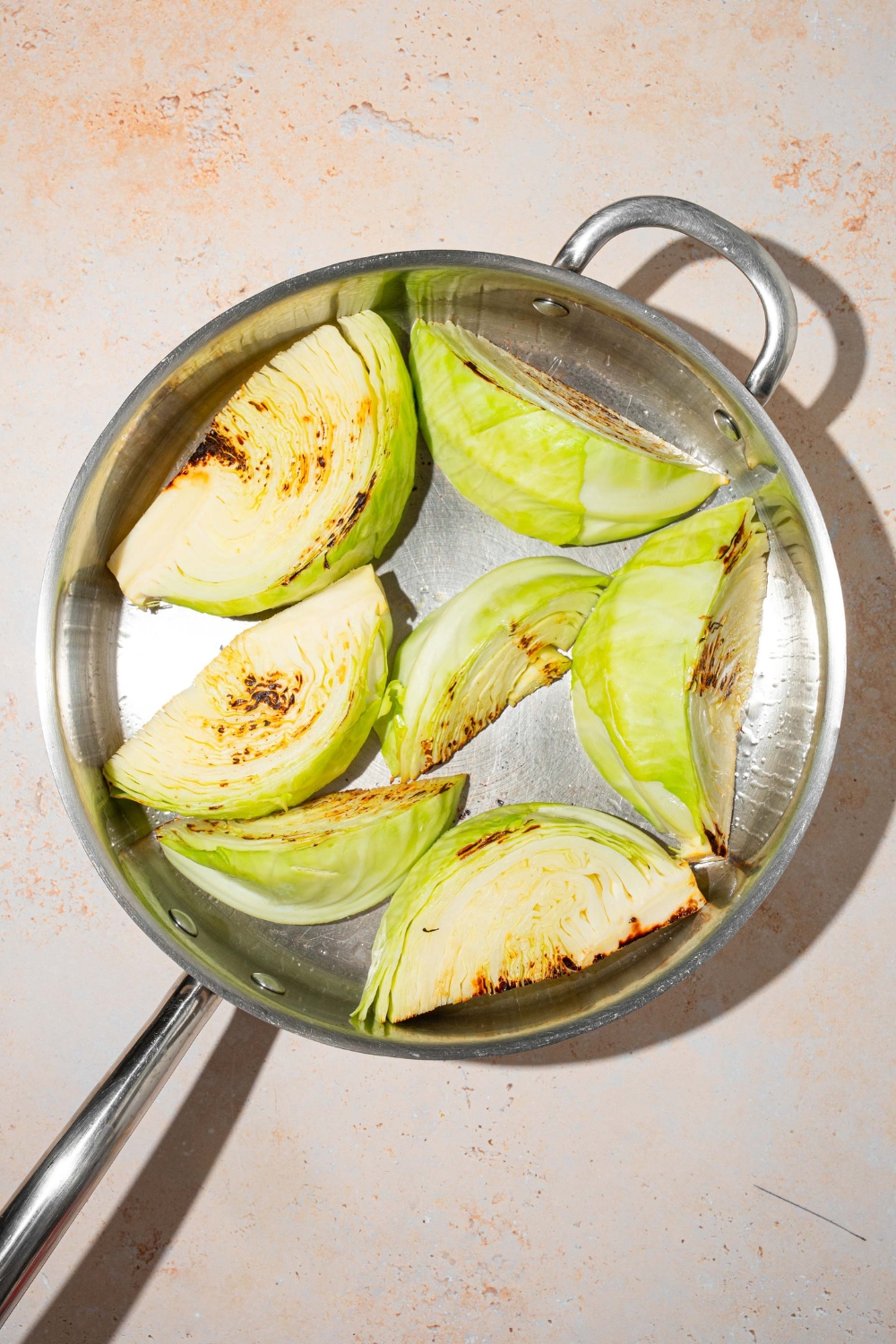 A skillet with several cabbage wedges sautéing in oil. The skillet is on a tan counter.