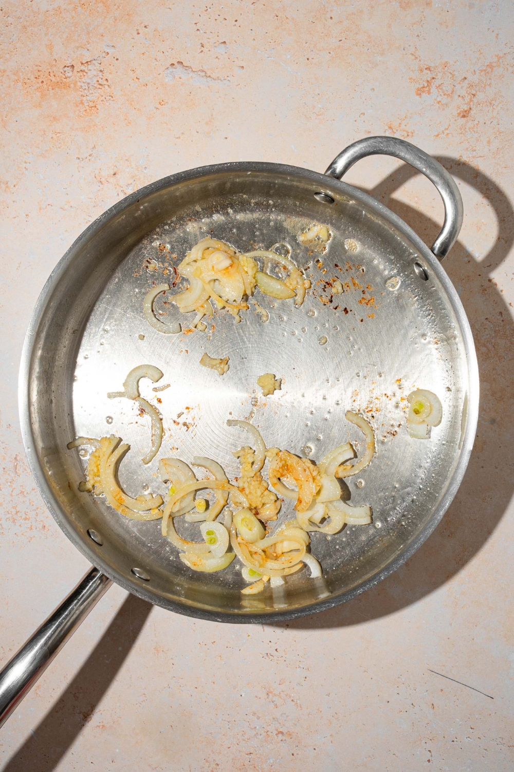 A skillet with sliced onion and garlic sautéing in oil. The skillet is on a tan counter.