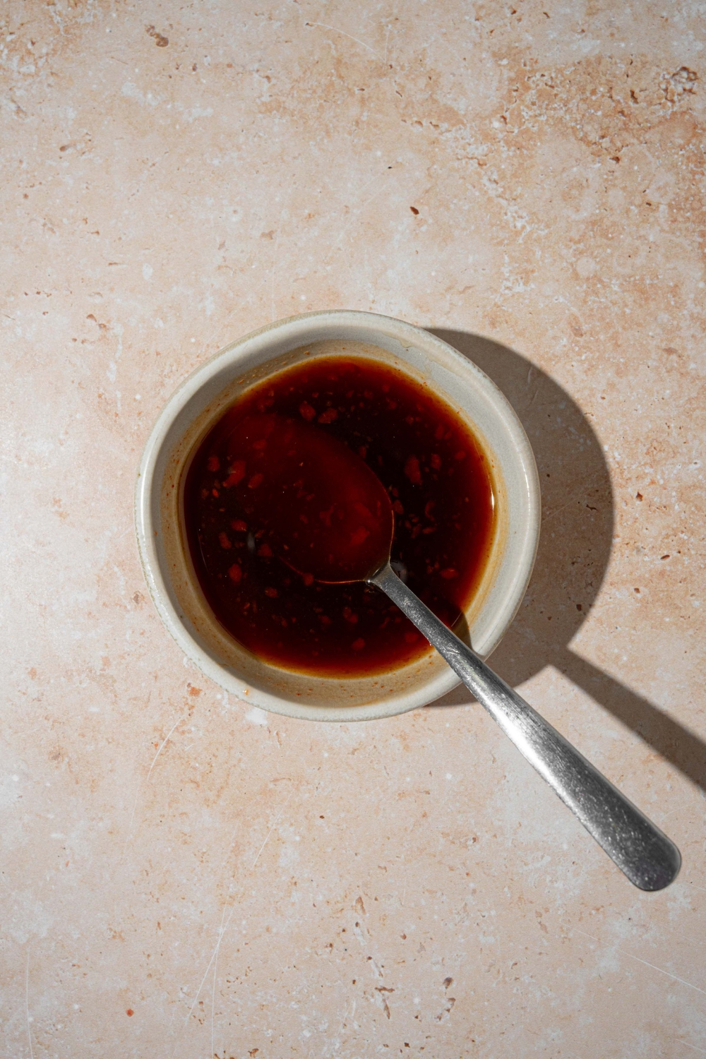 A small white bowl with a honey soy sauce mixture. There is a spoon in the bowl. The bowl is on a tan counter.