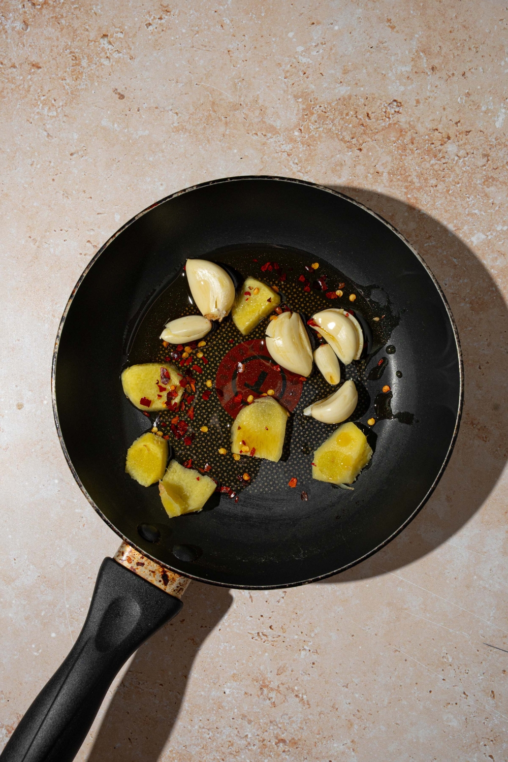 A skillet with smashed garlic, ginger, and chili flakes in oil. The skillet is on a tan counter.