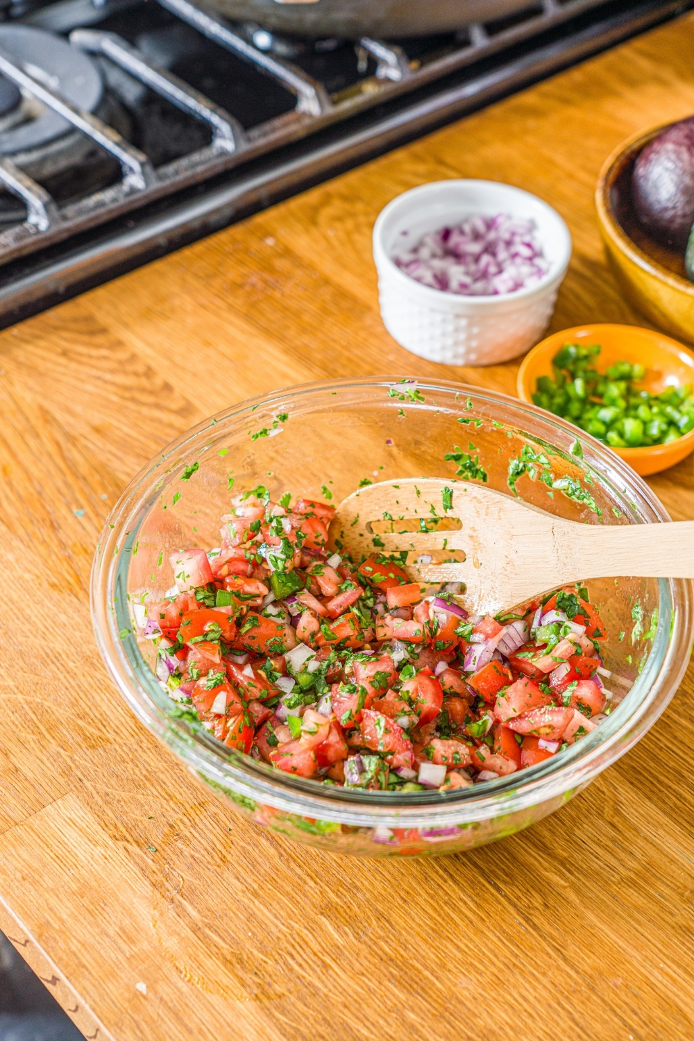A glass bowl with a wooden spoon mixing ingredients to make pico de gallo including tomatoes, red onion, jalapeño, and cilantro. The bowl is on a wooden counter.