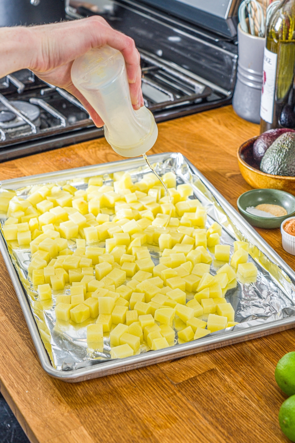 A baking sheet lined with foil with diced potatoes. A hand is squeezing oil over the potatoes.