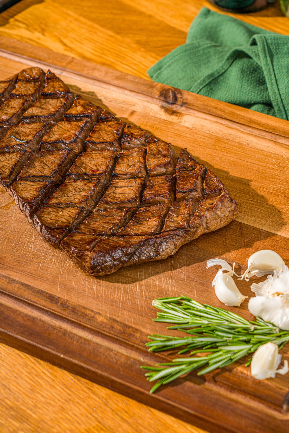 A wooden cutting board with a marinated and grilled London broil. The board is served with garlic and fresh rosemary.