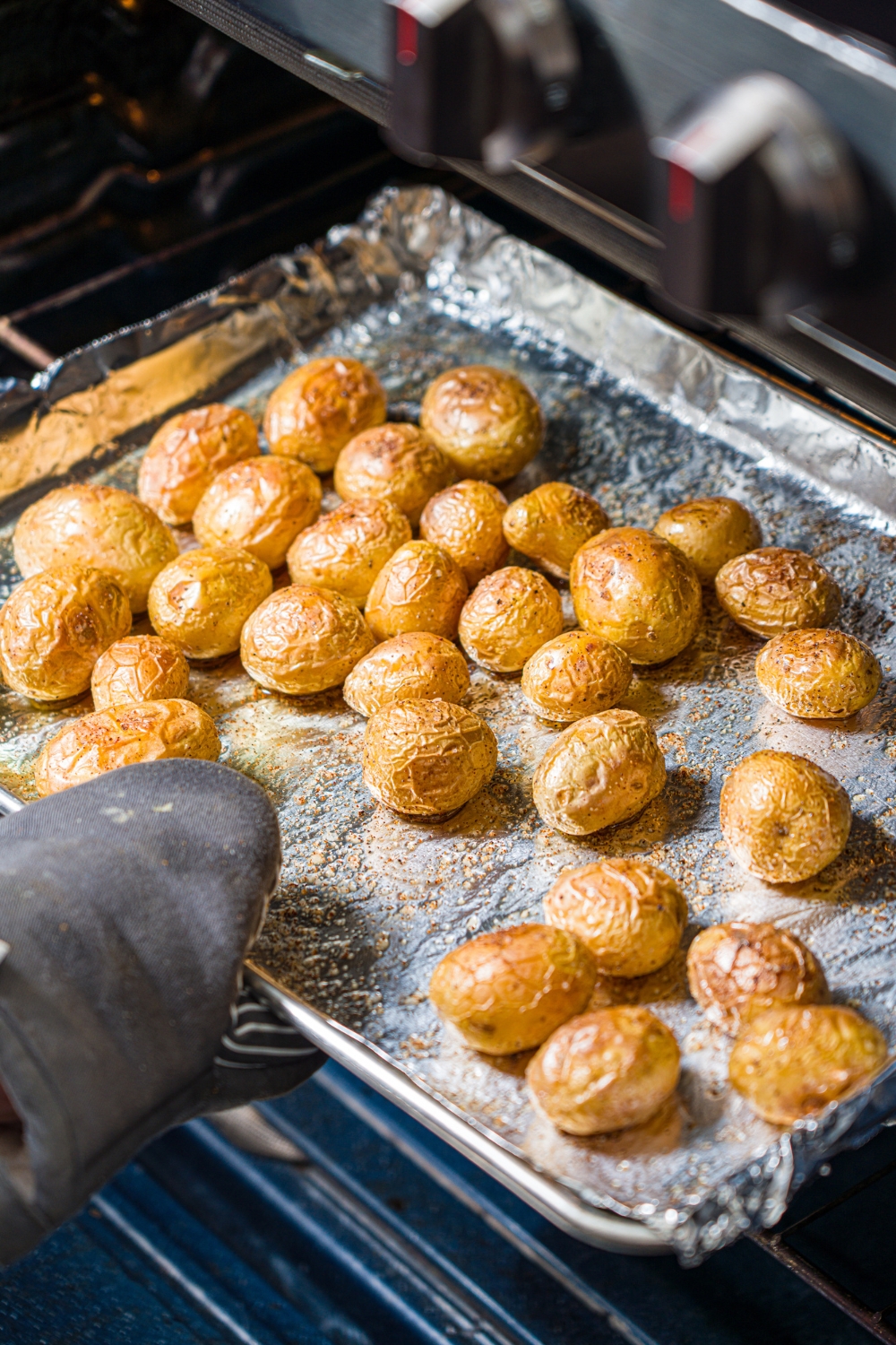 A baking sheet lined with foil with seasoned baked mini potatoes being pulled out of the oven.