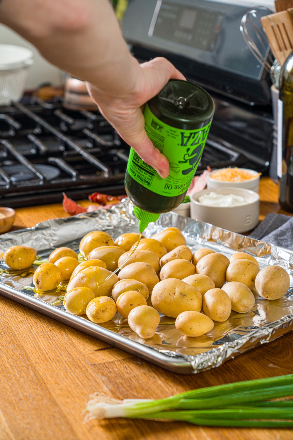A baking sheet lined with foil with mini potatoes being drizzled with oil. The sheet is on a wooden counter.