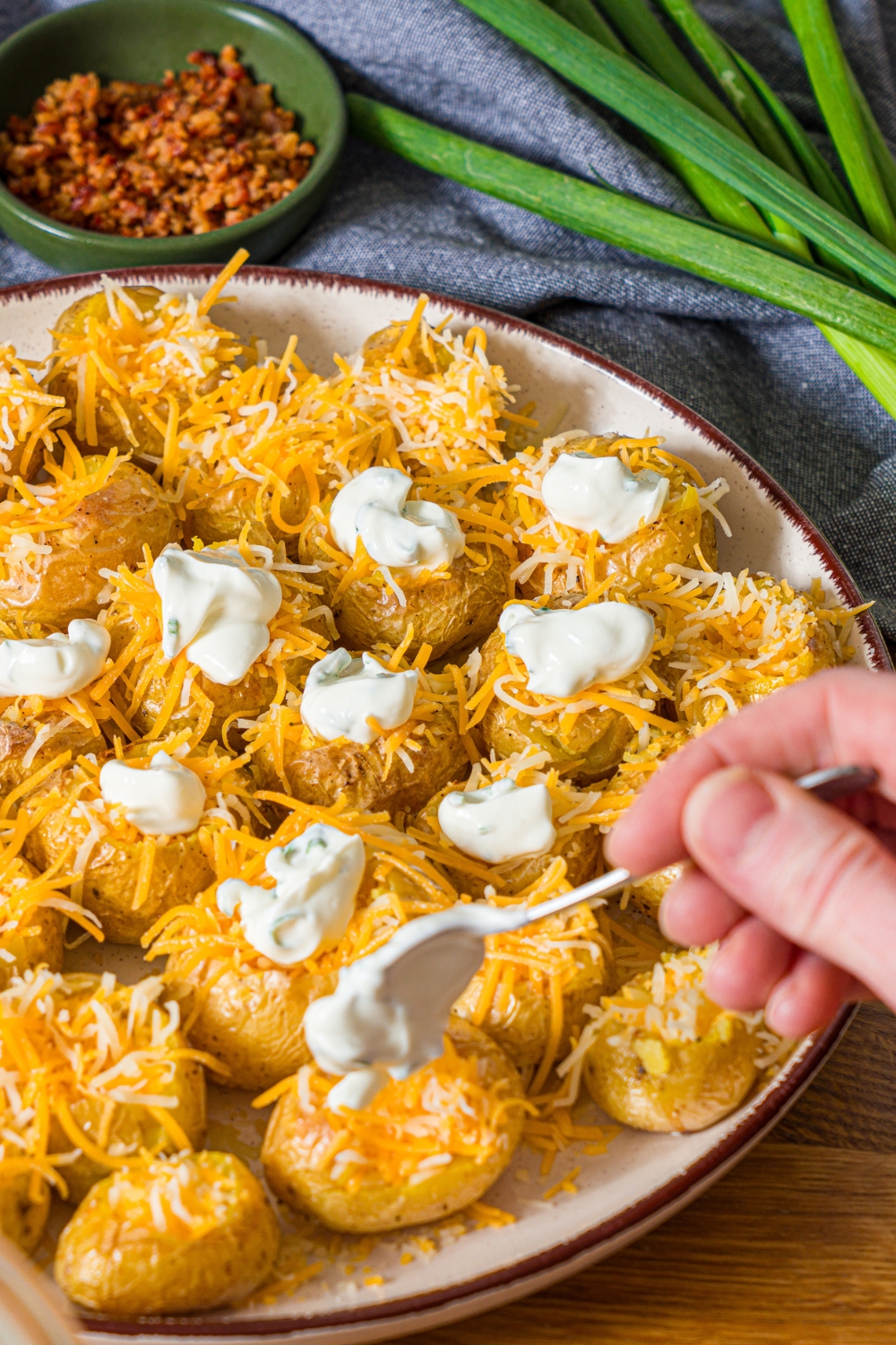 A white plate with mini baked potatoes being loaded with shredded cheese and sour cream. The plate is on a wooden counter.