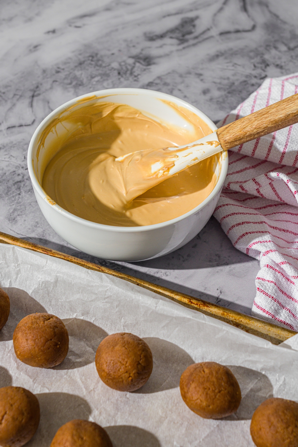 A white bowl of melted white chocolate with a spatula in the bowl. The bowl is on a marble counter with a baking sheet of truffles.