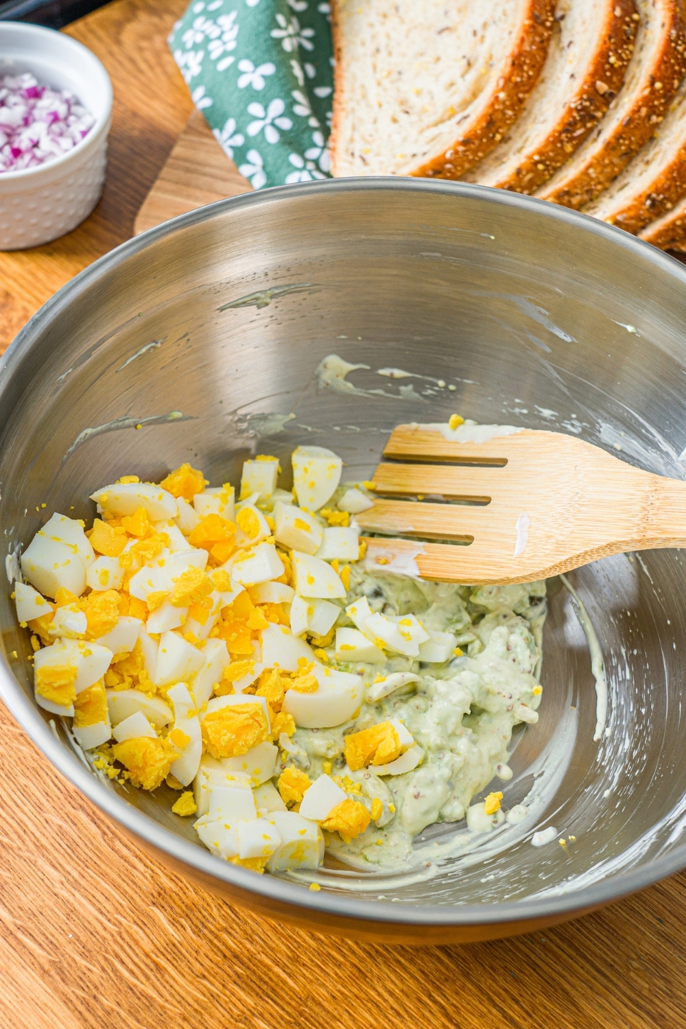 A large bowl with a wooden spoon mixing chopped egg with an avocado mixture. The bowl is on a wooden counter with sliced bread.