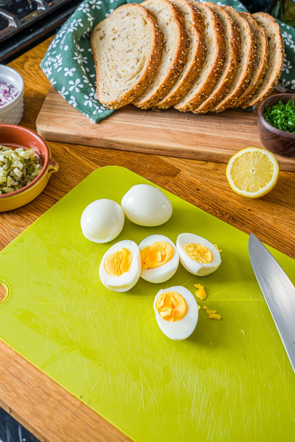 A green cutting board with several hard boiled eggs sliced in half. The board is on a wooden counter with a board of sliced bread.