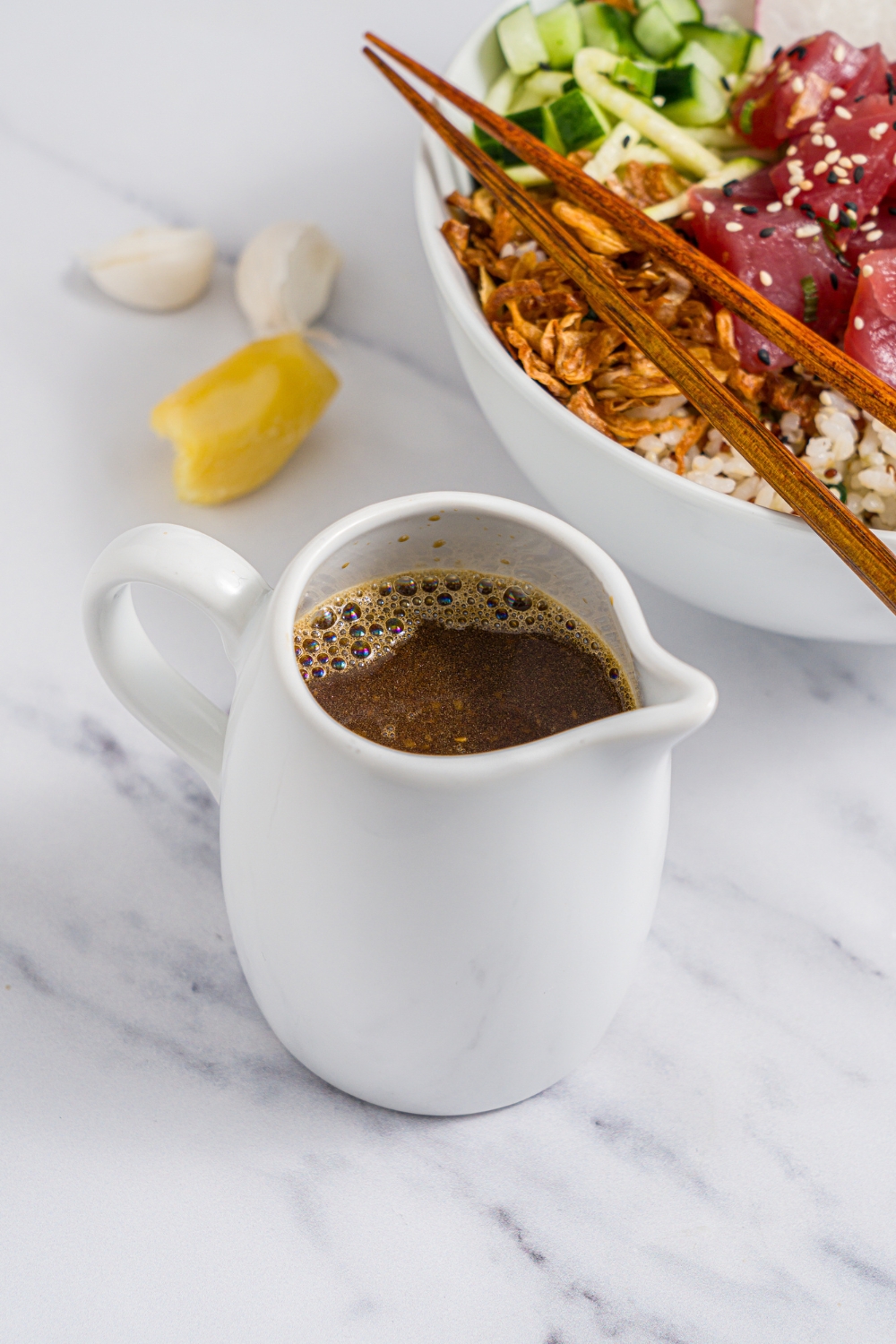 A small creamer filled with poke sauce. The creamer is on a white counter with a tuna poke bowl, garlic, and ginger.