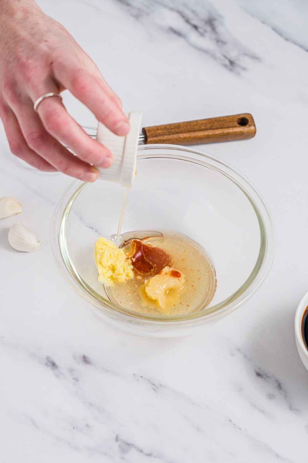 A glass bowl with ingredients being added to make poke sauce including ginger, sesame oil, rice vinegar, and seasonings. The bowl is on a marble counter.