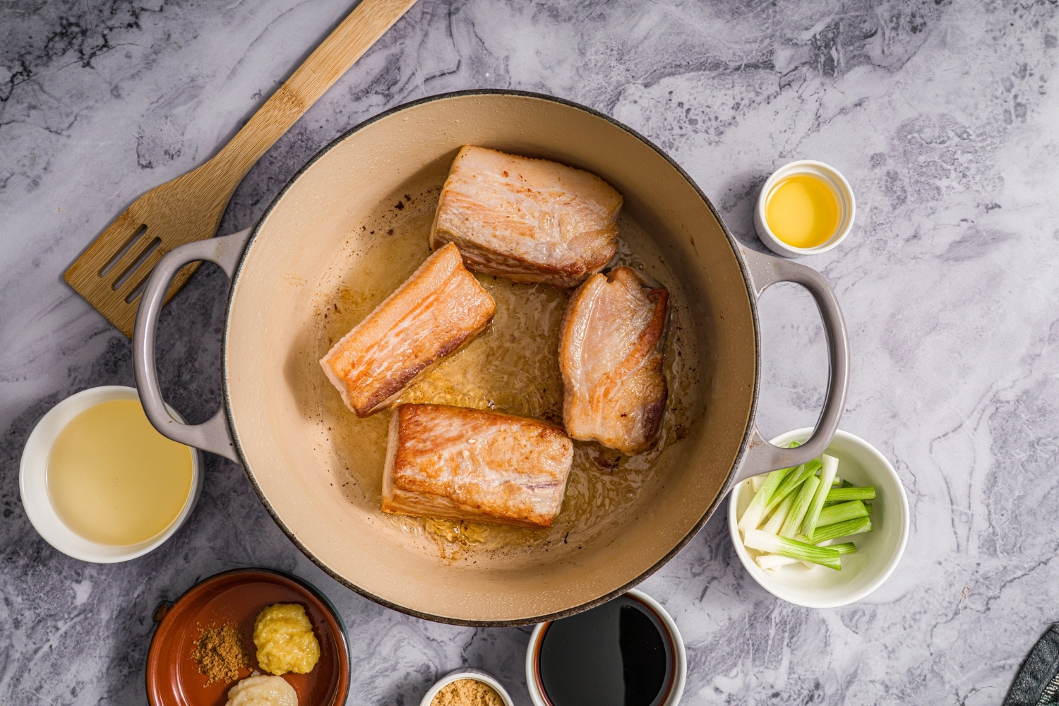 A dutch oven with pork belly searing in oil. The dutch oven is on a marble counter with small bowls of ingredients.