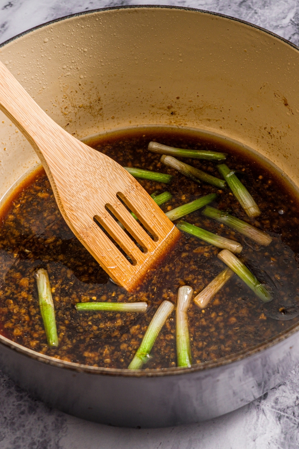 A dutch oven with a wooden spoon stirring a pork broth base. The pot is on a marble counter.