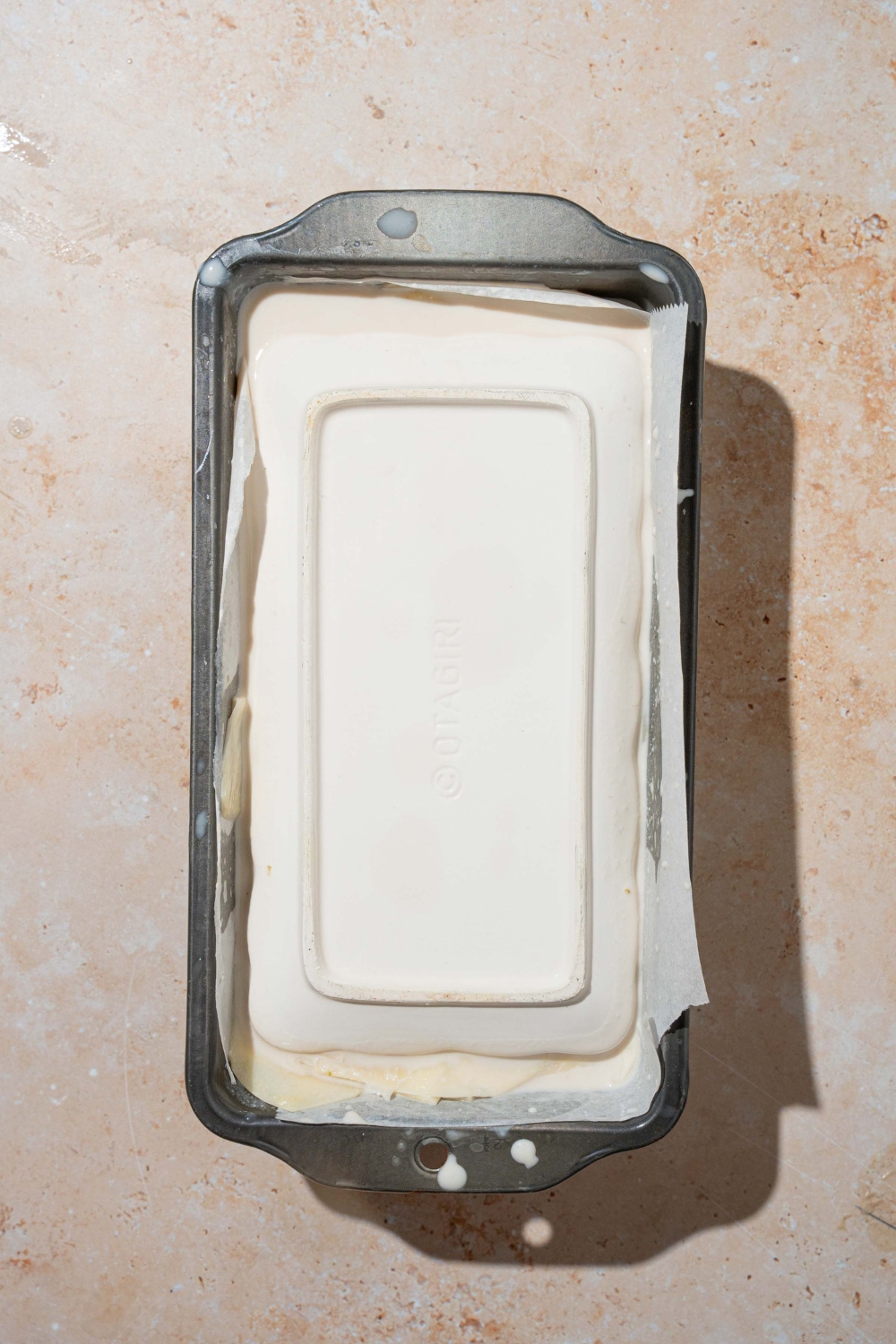 A loaf pan lined with parchment paper with thinly sliced potatoes soaked in cream and layered in the pan. A small plate is covering the loaf pan. The pan is on a tan counter.