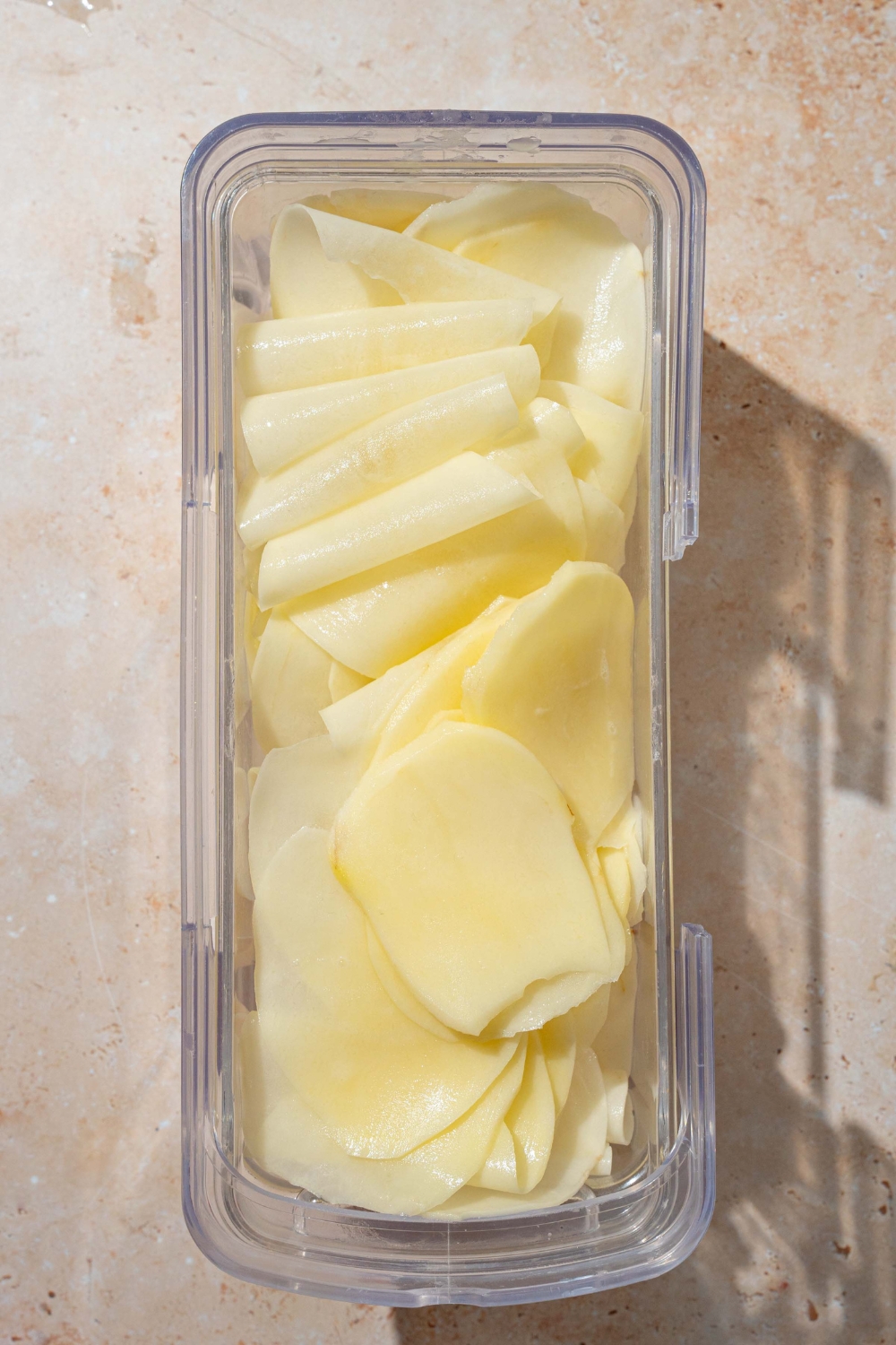 An airtight container filled with thinly sliced pieces of potato. The container is on a tan counter.